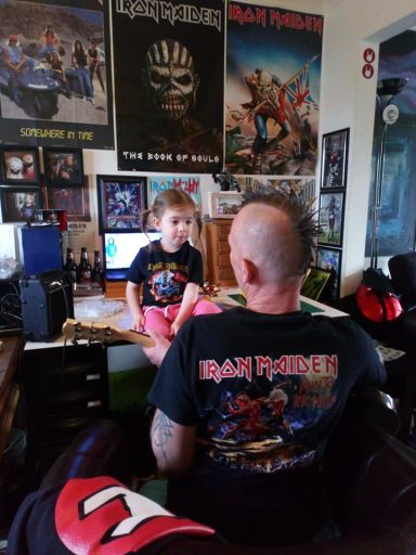 A child sits on a desk facing an adult with a mohawk, both wearing band t-shirts.