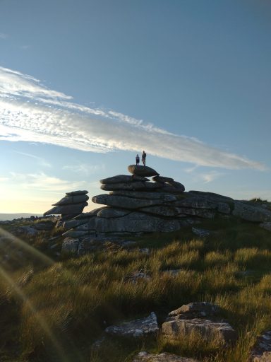 Two people stand on top of a large rocky outcrop against a clear blue sky.