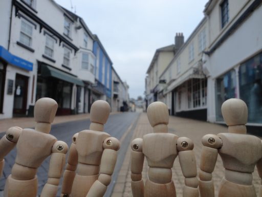 Four wooden mannequins stand on a quiet street lined with shops.