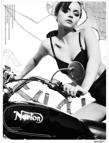 A woman poses on a classic motorcycle, wearing a black top against a graffiti backdrop.