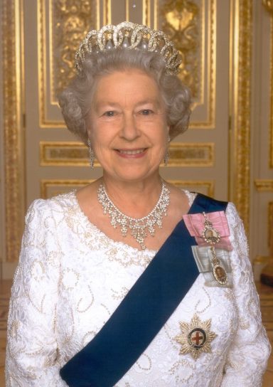 Elderly woman in a formal white gown with a tiara and silver jewellery, smiling warmly.