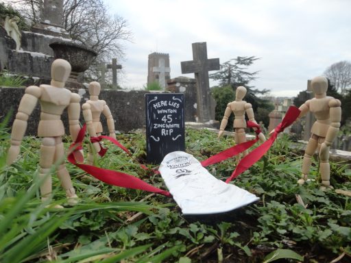 Wooden mannequins surround a gravestone tied with red ribbons in a graveyard setting.