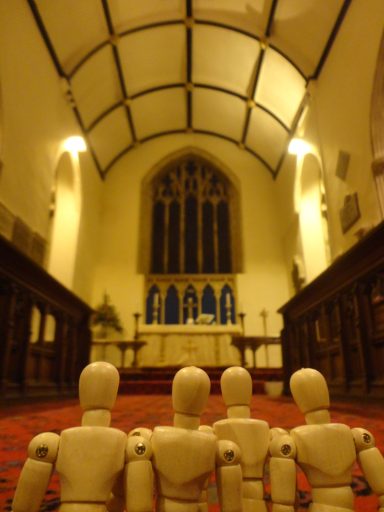 Four wooden mannequins face an altar in a church with high arched ceilings.