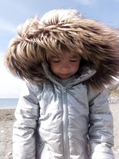 Child wearing a shiny silver coat with a fur-lined hood, standing on a beach.